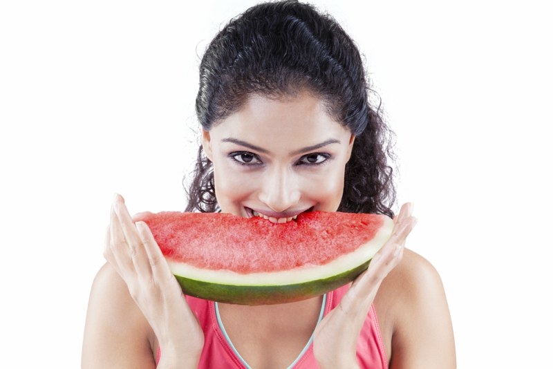 Beautiful Indian woman biting a big slice of sweet watermelon while smiling and looking at the camera, isolated on white background