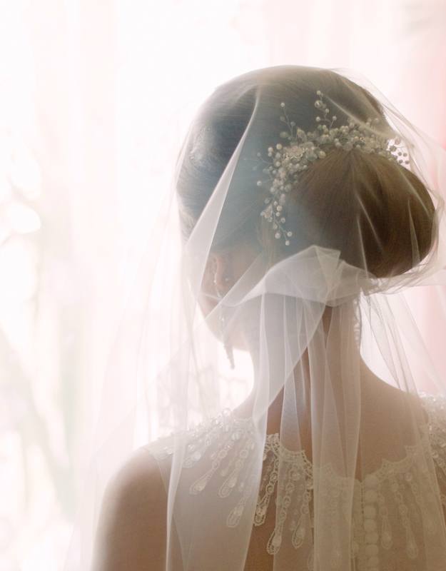 Tender brunette bride in white dress and veil standing back. Elegant hairstyle with stylish silver wedding accessories. Morning bridal preparation.