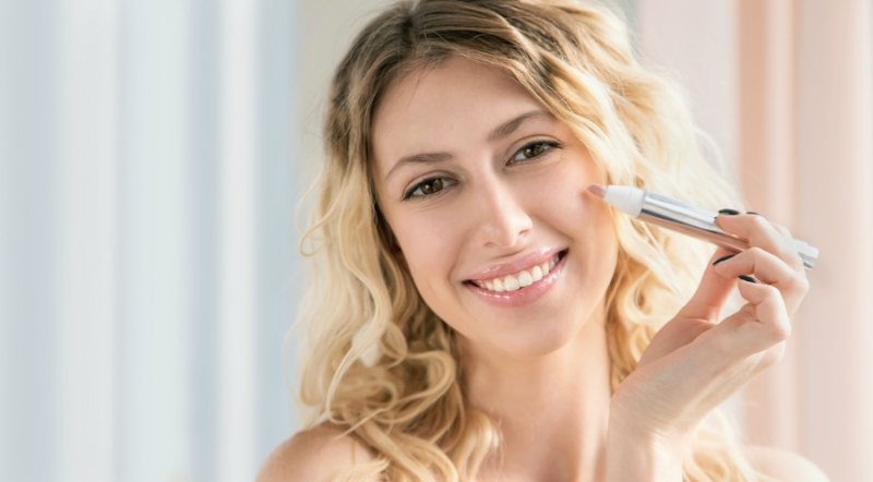 Young blonde woman applying make-up cosmetic product. Beautiful smiling girl with wavy hair use primer highlight concealer or foundation. Model presenting makeup or skincare output.