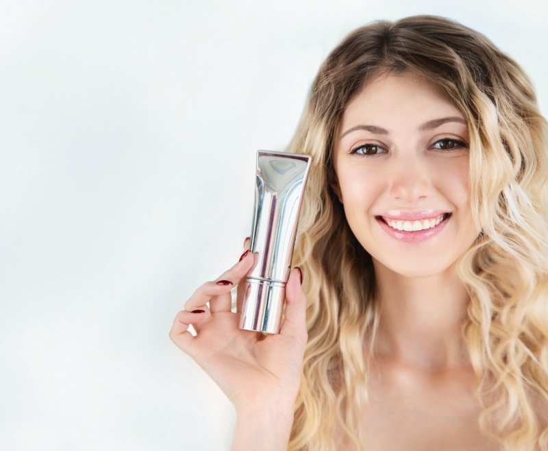 Young blonde woman presenting luxury cosmetic or make-up SPA products. Beautiful smiling girl with wavy hair holds of silver tube of moisturizer, oil, wellness, exfoliator, peeling cream in hand.