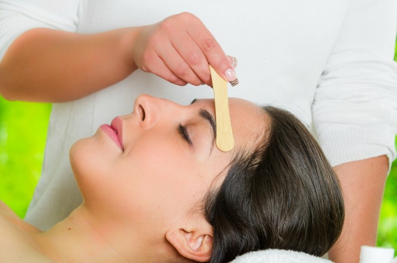 Closeup womans face receiving facial hair waxing treatment, hand using wooden stick to apply wax, beauty and fashion concept