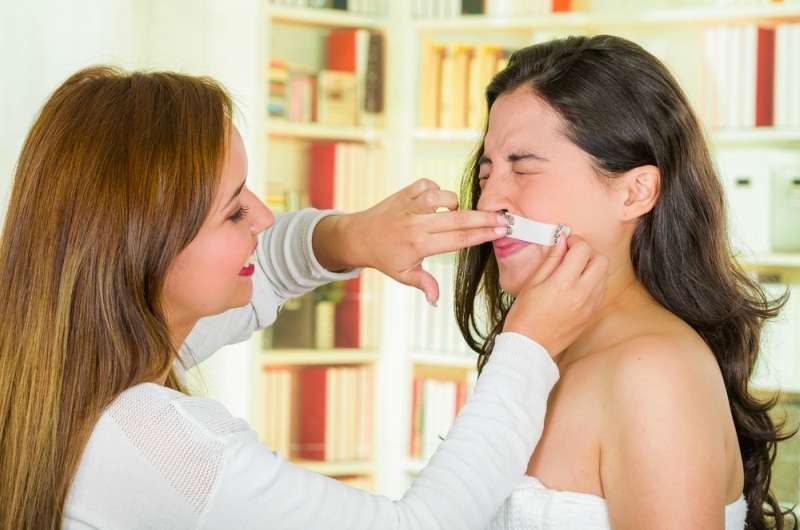 Cosmetologist applying wax on brunette clients upper lip, preparing for hair removal with painful facial expression