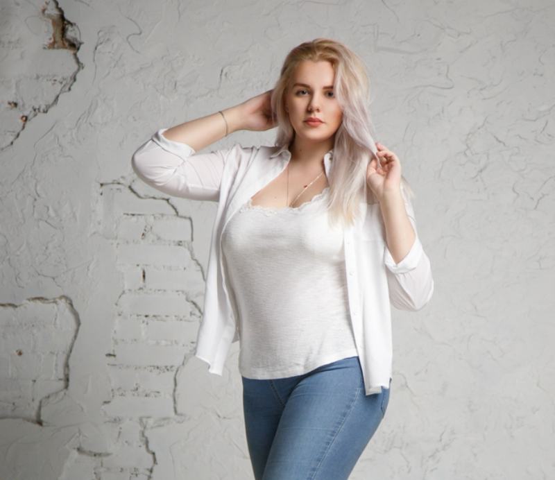 Portrait of a pretty big blonde in a white shirt on a gray background in full length.