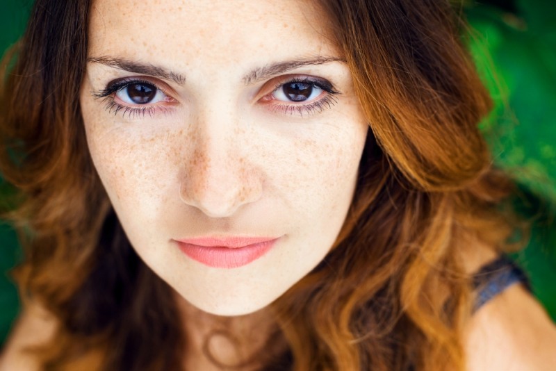 Portrait of young woman with the freckles and curly hair, against background of summer green park, green leaves. Natural beauty.