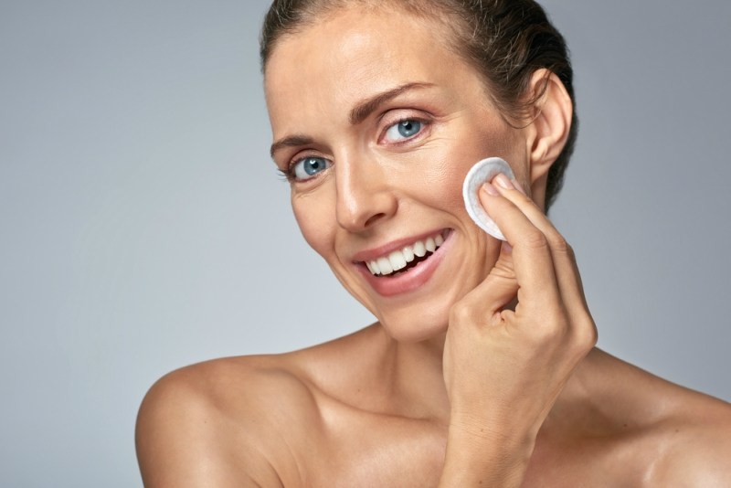 Smiling middle age woman wiping cleaning her face with cotton wool, wellness beauty skin care concept
