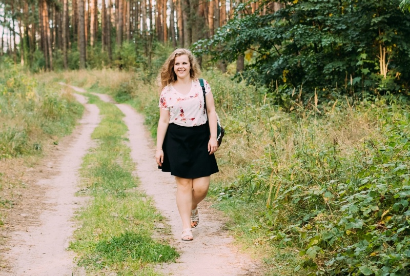 Young Pretty Plus Size Caucasian Happy Smiling Girl Woman In White T-Shirt And Black Short Skirt Walking Full-Length On Road In Summer Pine Forest