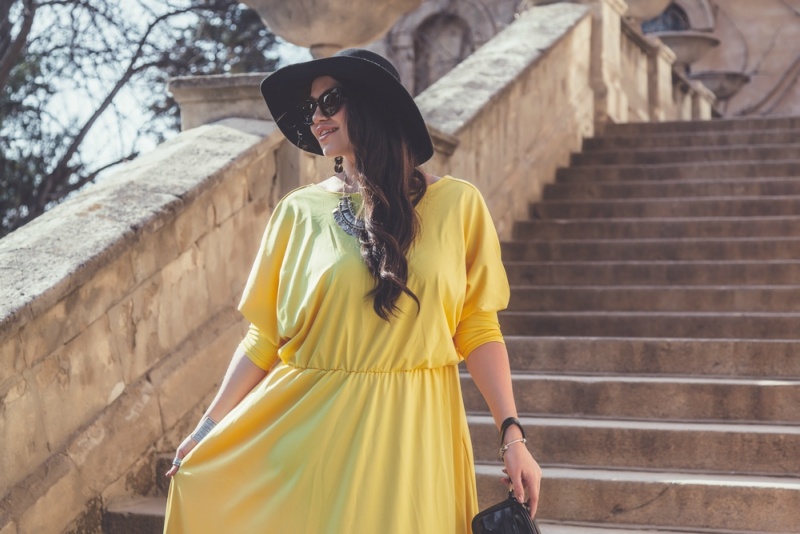 Young stylish woman wearing yellow maxi dress, black hat, sunglasses and handbag walking in the city street. Spring fashion outfit, elegant look. Plus size model.