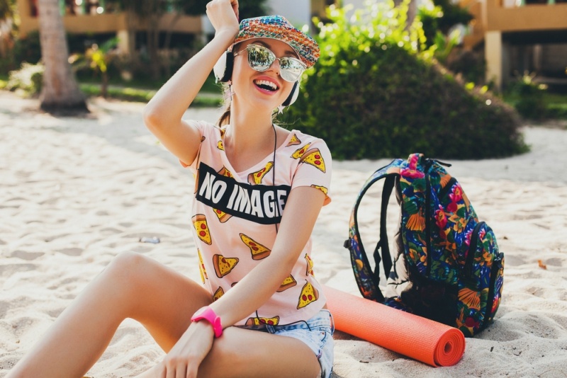 young beautiful woman walking on beach with yoga mat, listening to music on headphones, hipster sport swag style, denim shorts, t shirt, cap, sunglasses, sunny, summer weekend, cheerful