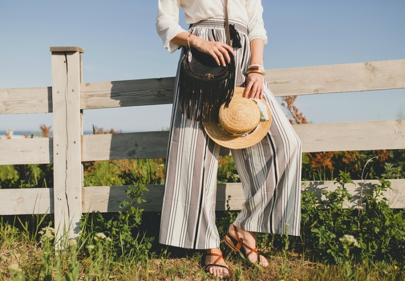 young stylish woman in countryside, holding a straw hat, summer, bohemian style outfit, sunglasses, bag, sandals, trousers