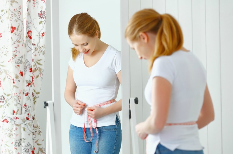 young woman measuring waist with tape in front of a mirror