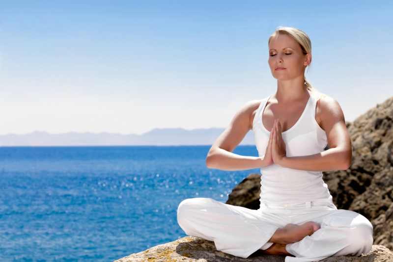 Beautiful positiveblond girl clothing in white sit at the seaside on the rock and meditating in yoga pose