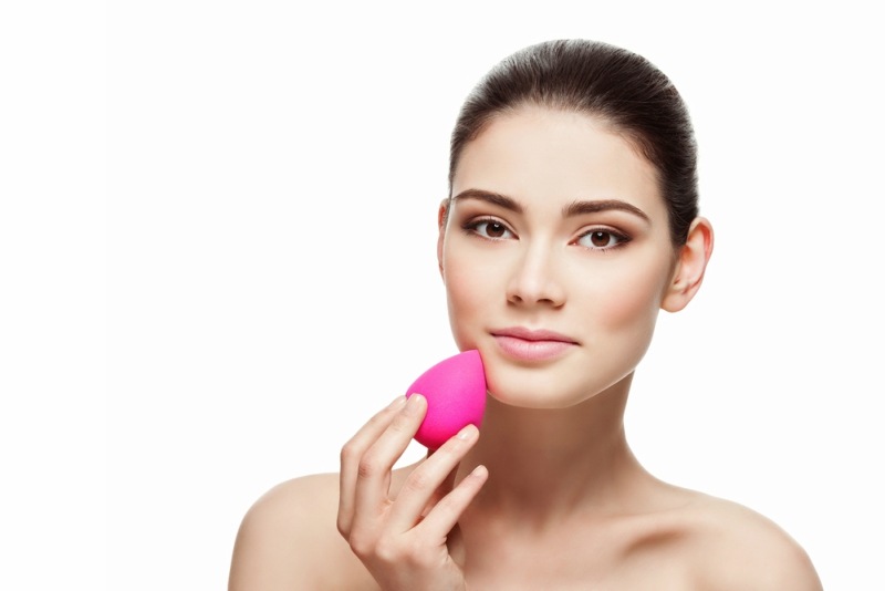 Beautiful young woman applying makeup using beauty blender sponge. Isolated over white background