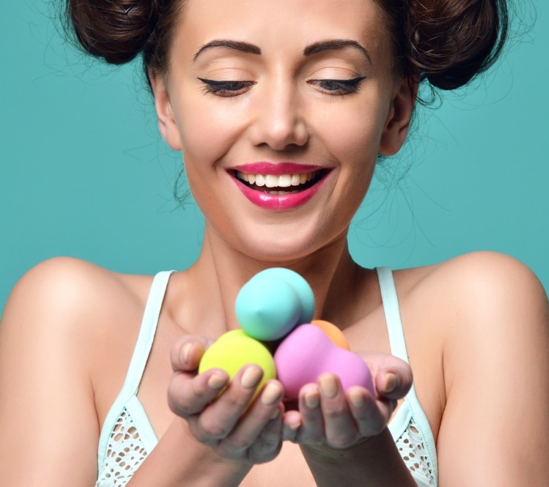 Happy surprised woman hold colorful sponge for applying foundation for make-up on face skin cares on blue mint background