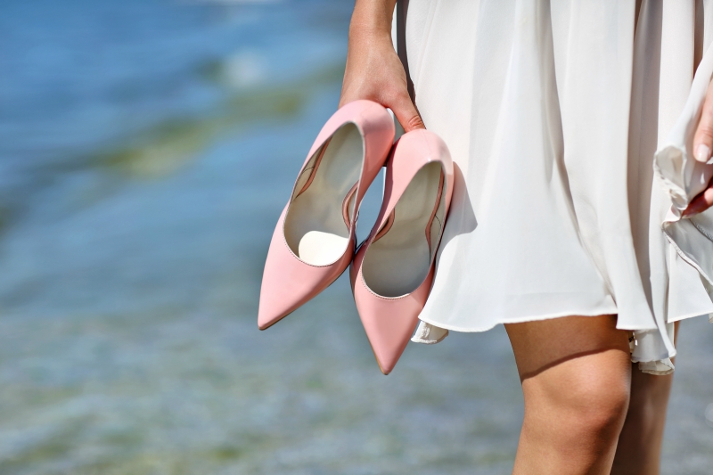woman holding pink stiletto heels on the beach walking in white dress